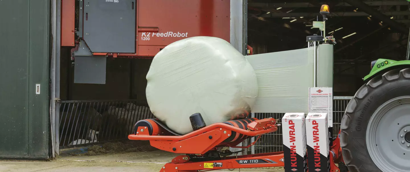 RW 1110 wrapping a round bale in front of a barn with a Feed robot at the background.